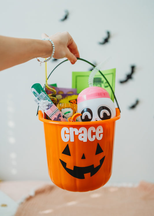 Orange Halloween bucket with 'Grace' printed on it, filled with treats and a ghost toy, against a white background.