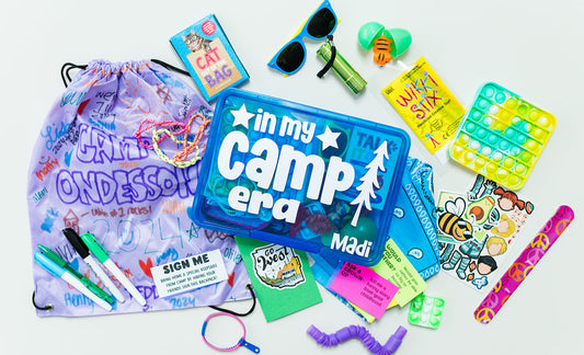 A collection of items for a summer camp care package, including a cinch-string backpack, permanent markers, a card game, sunglasses, a fidget pop tube, zipper bracelet, bendy snake, slap bracelet, activity cards, a sleep mask, a notebook with stickers, a mini pop it keychain, and a mochi squishy.