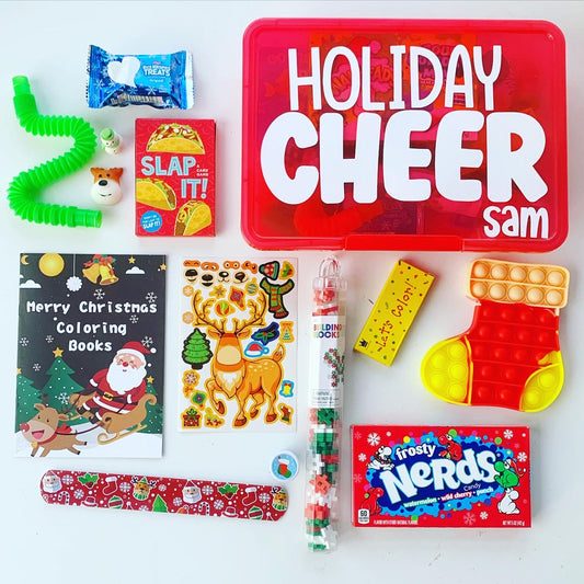 A red plastic box with 'HOLIDAY CHEER' text containing various holiday-themed items for kids, including a coloring book, building blocks, crayons, a sticker sheet, a slap bracelet, a pop tube fidget toy, a card game, sprinkled rice krispie treat, and two mochi squishy fidget toys.