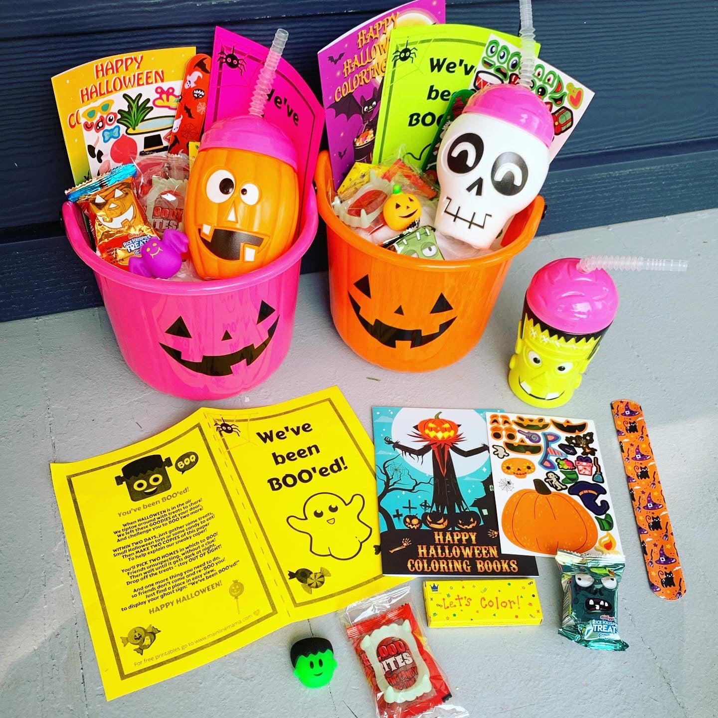 Two pink Halloween-themed baskets filled with various treats and items, including plastic fangs, a Halloween cup, straw, eyeball candy, mochi squishy, face sticker sheet, slap bracelet, mini coloring book, crayons, and a Rice Krispie Treat. There is also a 'You've been BOOed' instruction sign and tissue paper for stuffing.