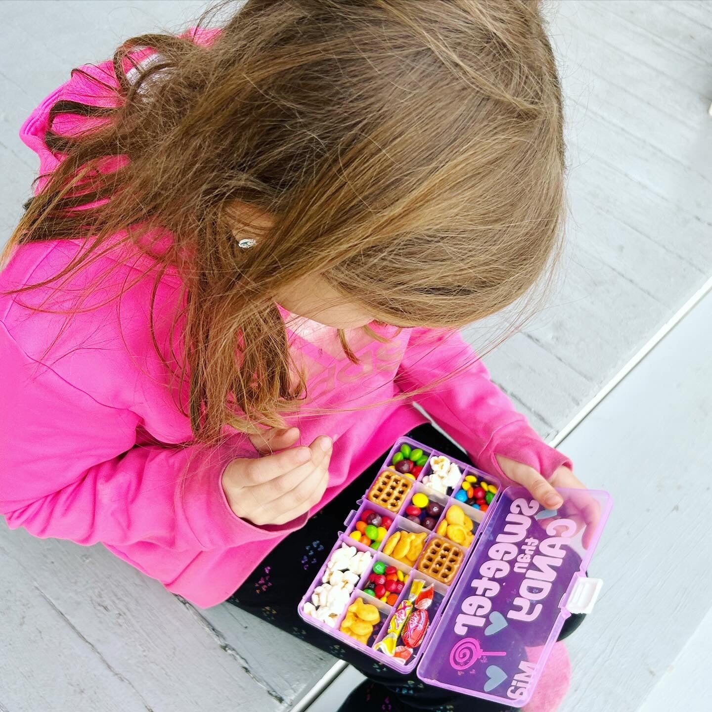 Young girl looking down at a pink snack box open in her lap with pretzels, skittles, m and ms, popcorn, candy, goldfish crackers inside. Top of snack box says sweeter than candy with name Mia.