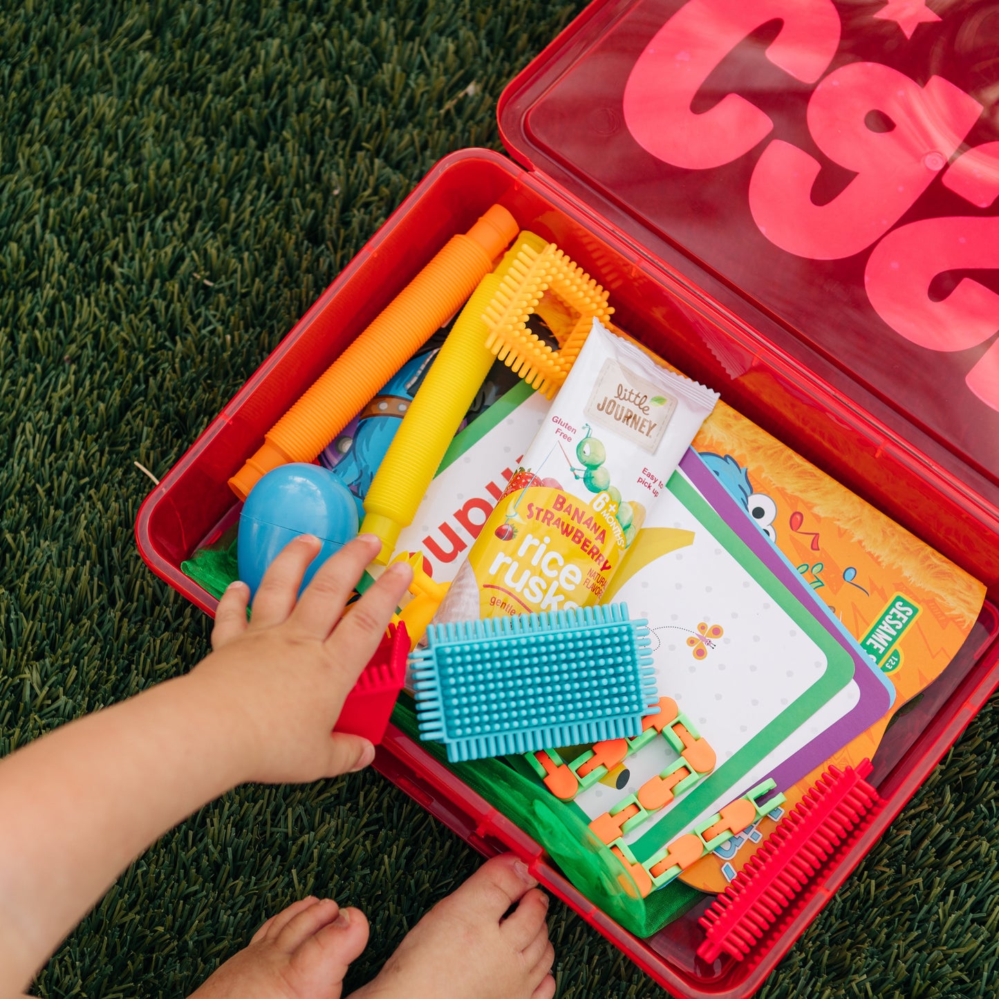 A child's hands reaching into a red busy box filled with various colorful toys and activities suitable for kids, such as coloring books, Wikki Stix, and plastic animals.