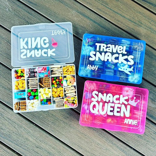 3 travel snack boxes. Clear box open with snacks inside each compartment. Goldfish crackers, skittles, tootsie rolls, pretzels, animal crackers, candy inside. Blue box with travel snack, name Abby and airplane. Pink Snack Queen box with crown.