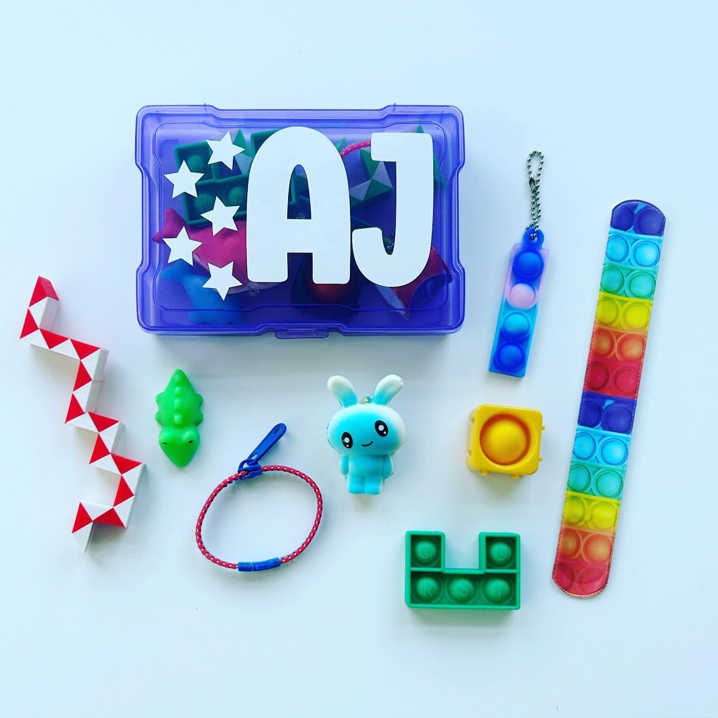 A personalized fidget storage box with various colorful fidget toys displayed in front, including a snake fidget, a squishy, a pop it line keychain, and other small toys. The box is purple with a name written on it.