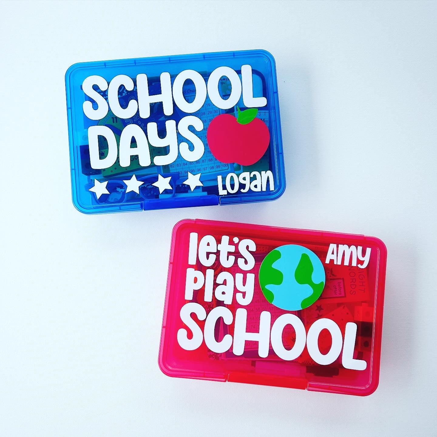 Two pretend school play sets with 'School Days' and 'Let's Play School' text, each with a different color background. One set is blue with a star applique and the name 'Lola' on the front, and the other is red with a 'School' text and a globe applique.