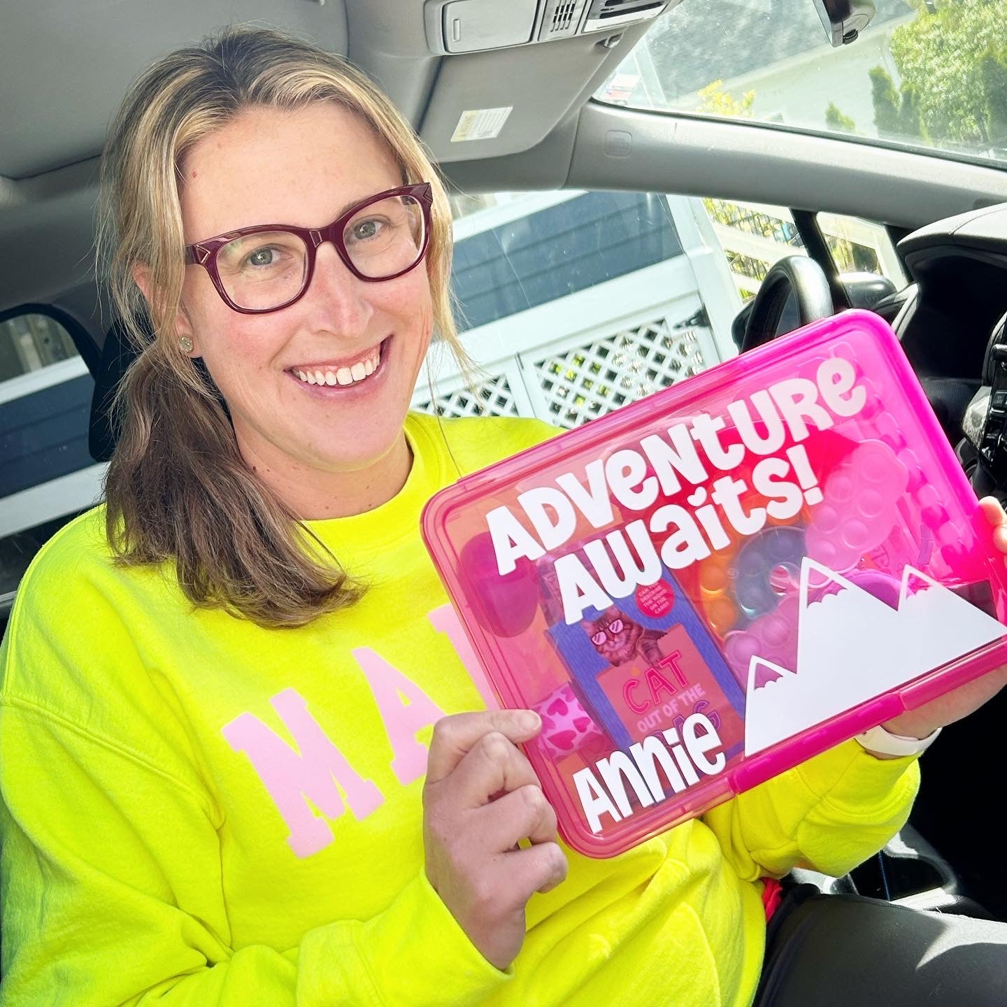 A person holding a pink busy box labeled 'ADVENTURE AWAITS' with various travel-friendly activities inside.