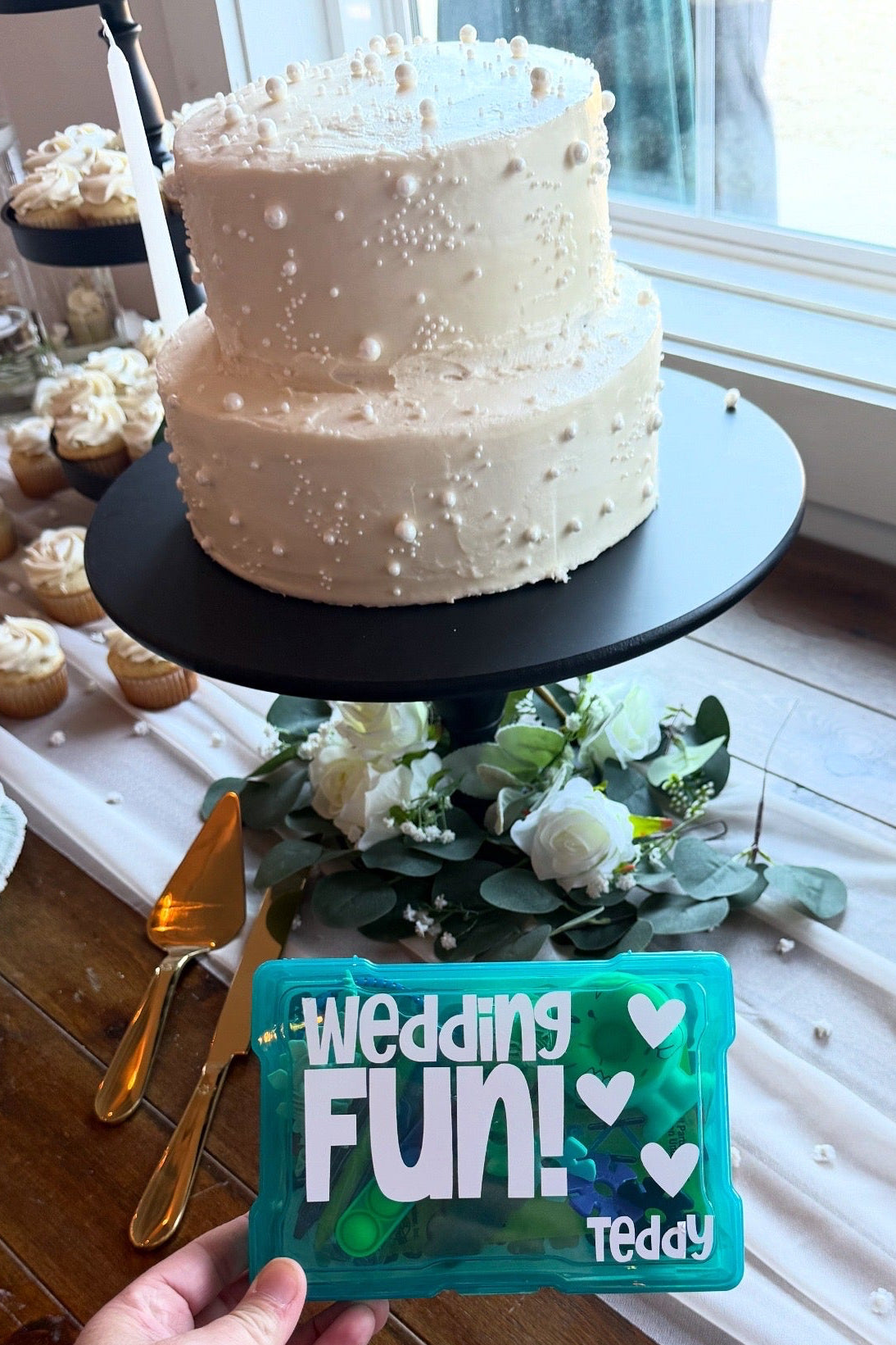 Two-tiered white wedding cake with decorative elements on a table, held by a person with a 'Wedding Fun! Teddy' sign.