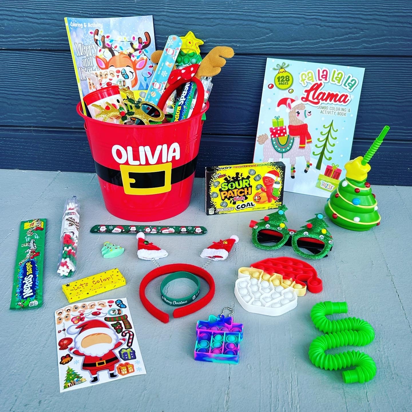 A red Christmas gift basket filled with various children's toys and treats, including a Santa hat, fidget toys, a coloring book, and candy.