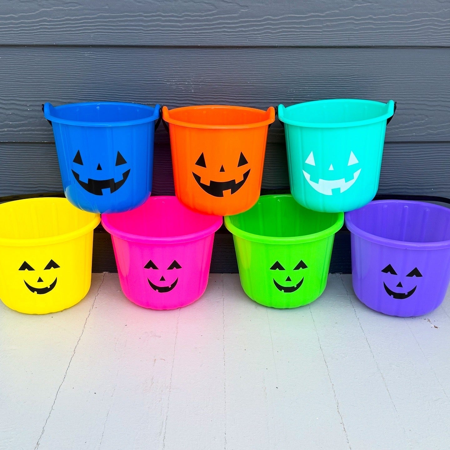 Colorful Halloween buckets with jack-o'-lantern faces against a gray wall.