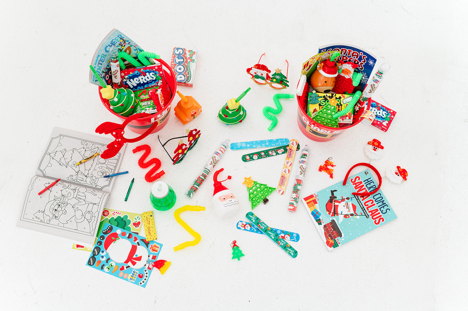2 red Christmas buckets filled with Christmas and holiday themed goodies for kids. Surrounding the buckets are Christmas coloring books, activities, fidgets, novelty glasses and headbands, slap bracelets, stickers, cups and more.