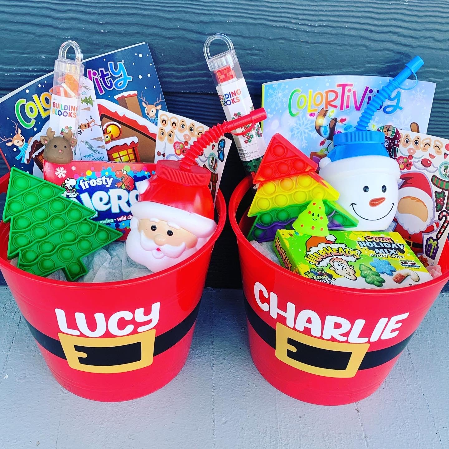 Two red Santa buckets serving as gift baskets, one labeled 'Lucy' and the other 'Charlie', filled with various Christmas-themed toys and items.