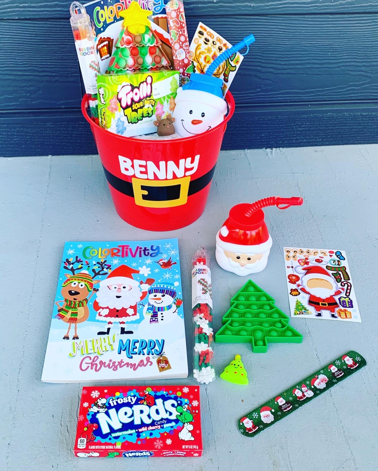 Christmas bucket with the name Benny on it which is filled with candy, pop it, coloring book, stickers and more. Contents are also laid out in front of the bucket.