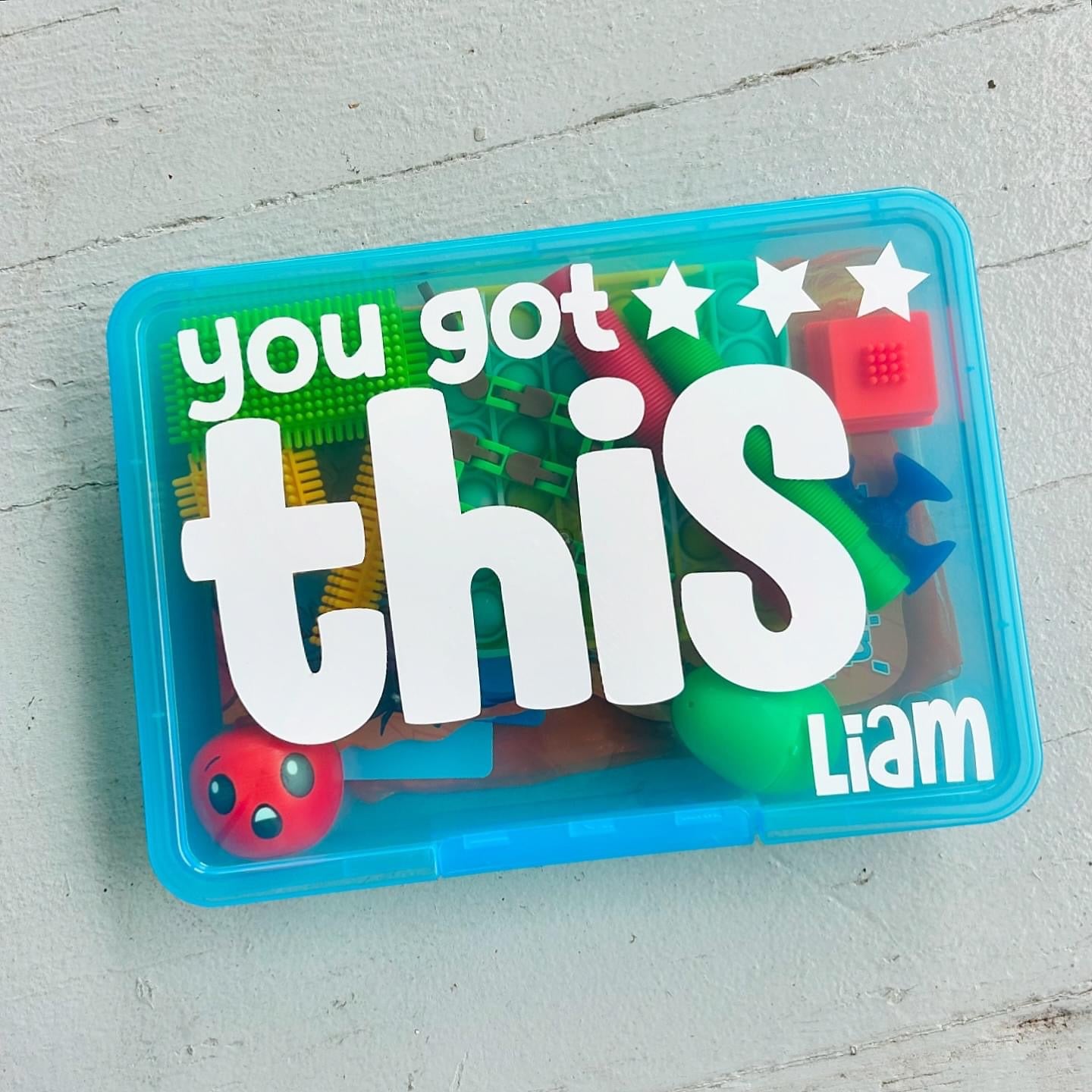 A get well soon care package for kids, containing various activities and toys, sealed in a blue container with a transparent lid, labeled with a name and get well messages.