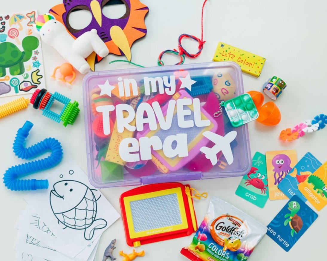 A variety of colorful children's toys and activities spread out, including a magnadoodle board, coloring pages, stickers, and other small items with a 'Travel' themed busy box in the center.