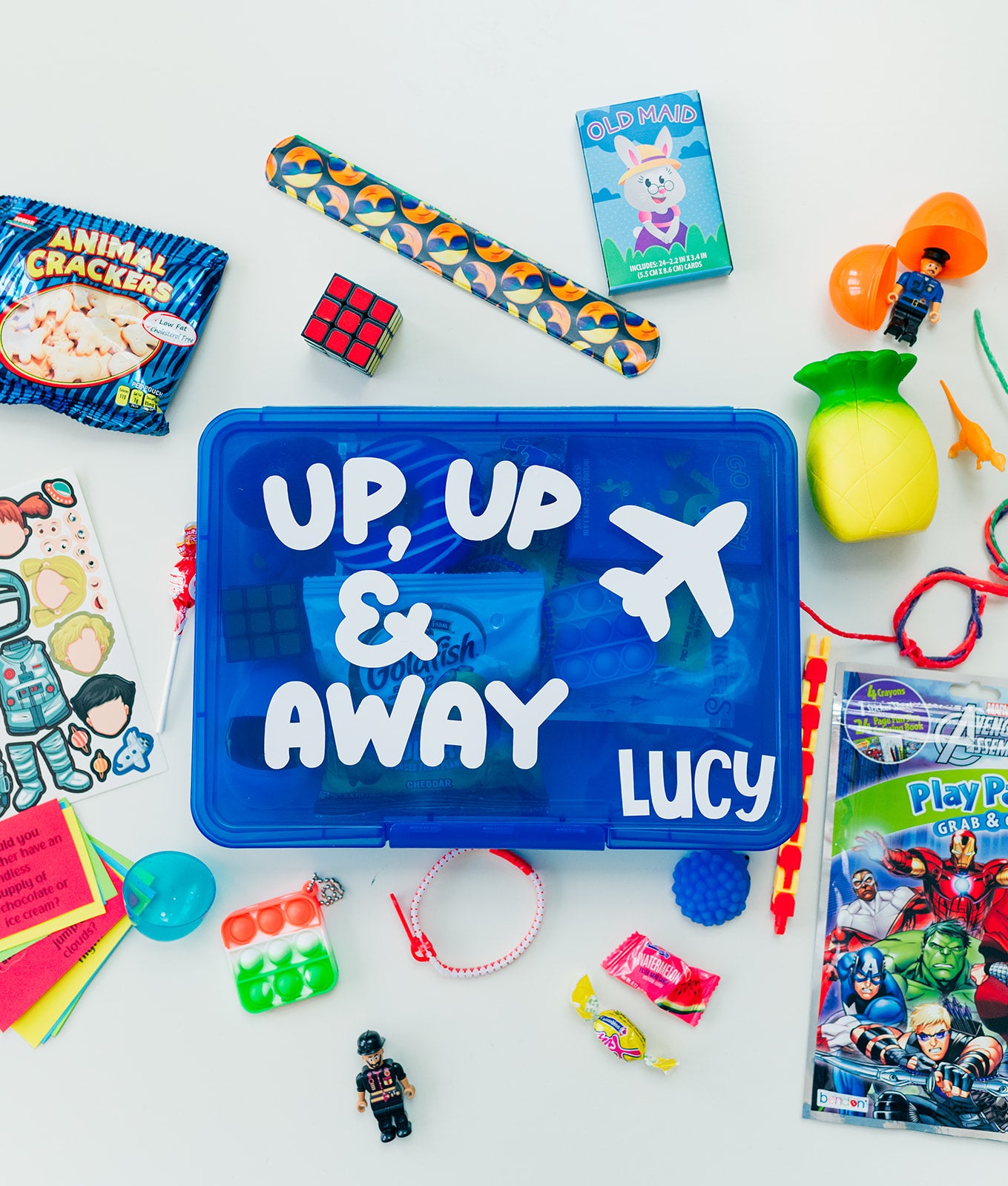 A travel busy box for kids with various activities displayed, including coloring books, stickers, mini figures, and more, with a blue lid that has 'UP, UP & AWAY LUCY' written on it.