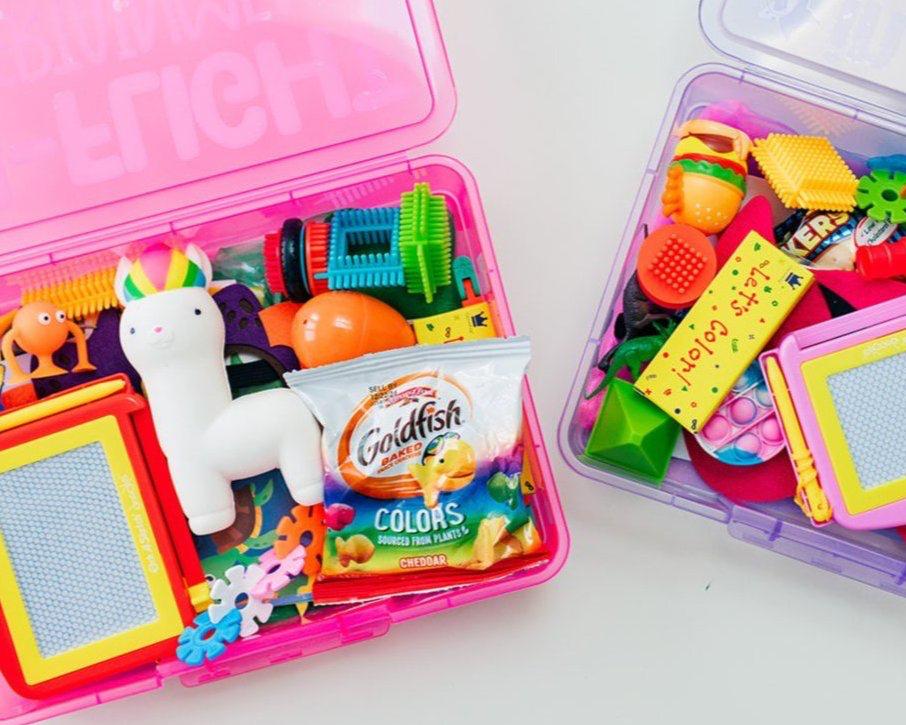 A colorful travel busy box for children containing a variety of small toys and activities such as a mini magna doodle, superhero mask, wikki stix, a set of make a face stickers, bristle blocks, plastic animals, a pop it keychain, slap bracelet, crayons, coloring pages, a slow rising squishy toy, suction cup toy, flashcards, and a brain flake STEM toy, all visible in an open pink zippered case.