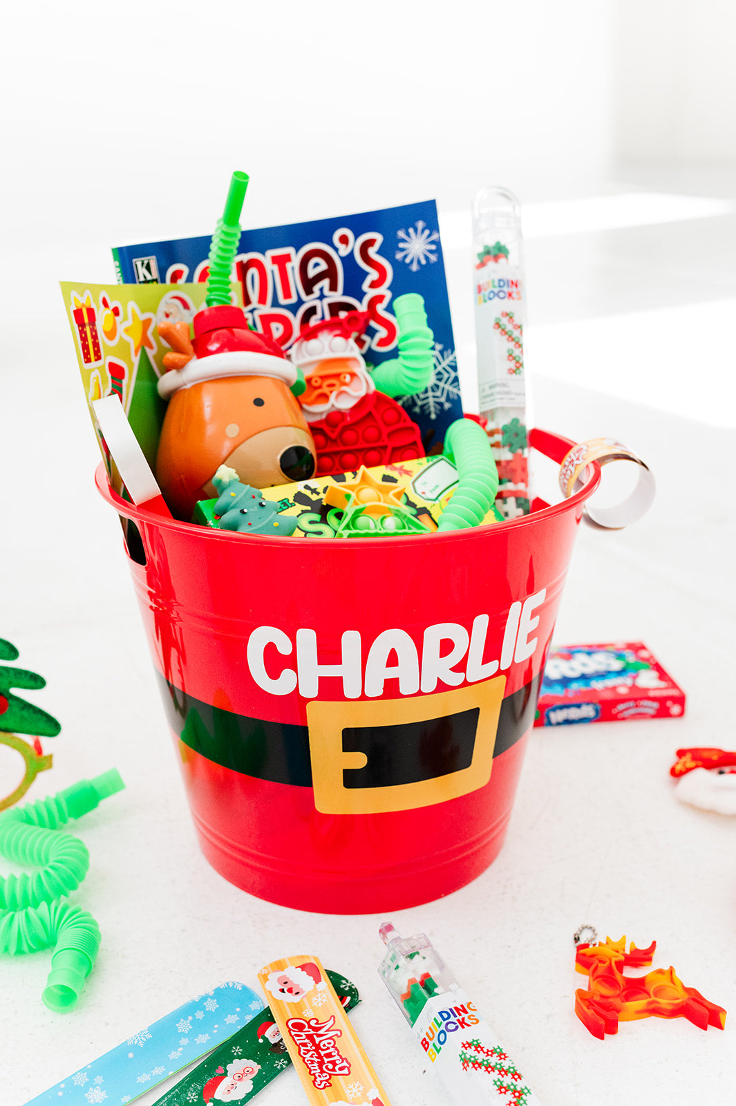 Red bucket labeled 'Charlie' filled with Christmas-themed toys and treats on a white background
