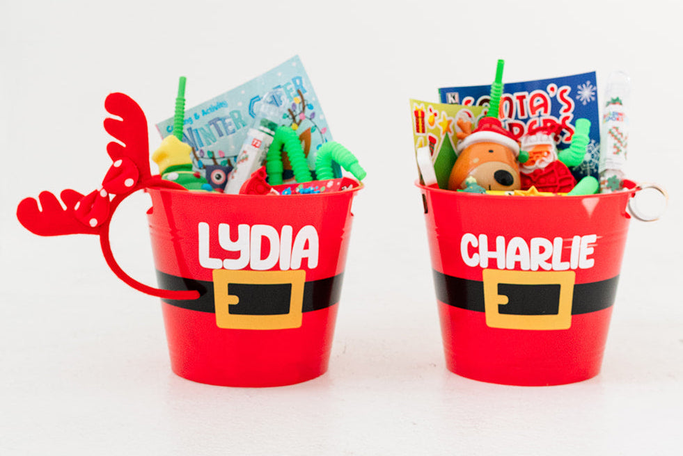 Two red Santa-themed buckets labeled 'Lydia' and 'Charlie' filled with toys on a white background.