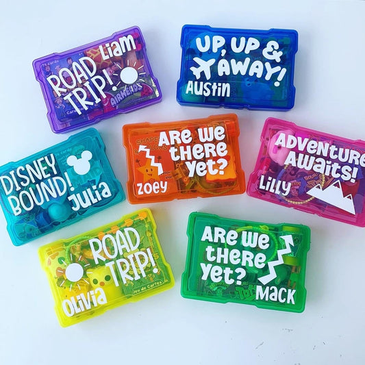 A collection of colorful mini busy boxes labeled with various names and travel-themed phrases such as 'ROAD TRIP!' and 'Are we there yet?'.