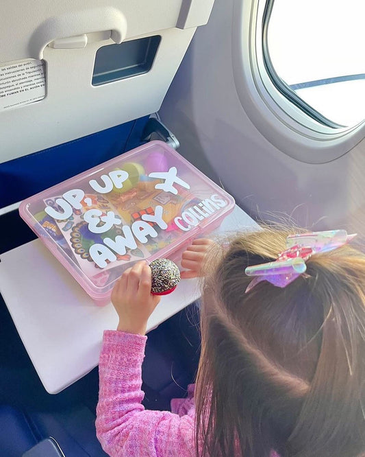 Young girl on an airplane with her airplane activity busy box on the airplane tray table. Light pink box with "up, up and away" and the name "Collins"