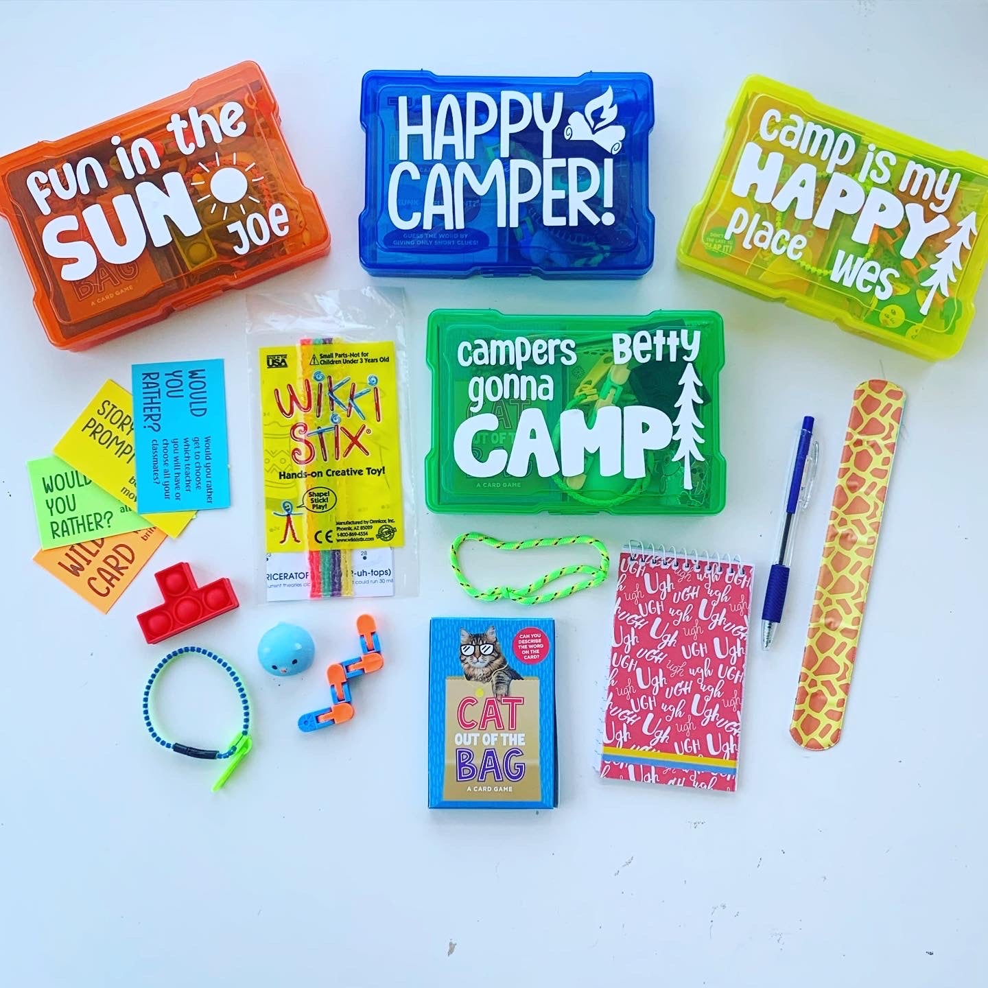 A collection of children's summer camp care package items including a notebook, zipper bracelet, Wikki Stix, a cat-themed book, a blue zippered pouch, and other small toys.