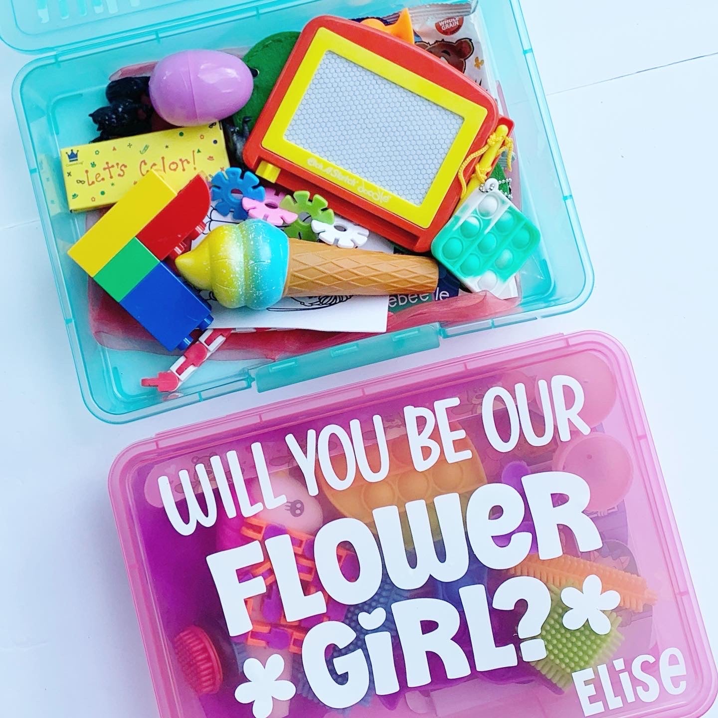 2 busy boxes. Turquoise blue one is open showing activities inside including legos, squishy, magna doodle, crayons, fidgets, plastic easter egg. Pink box is closed with will you be our flower girl and name Elise on the front.