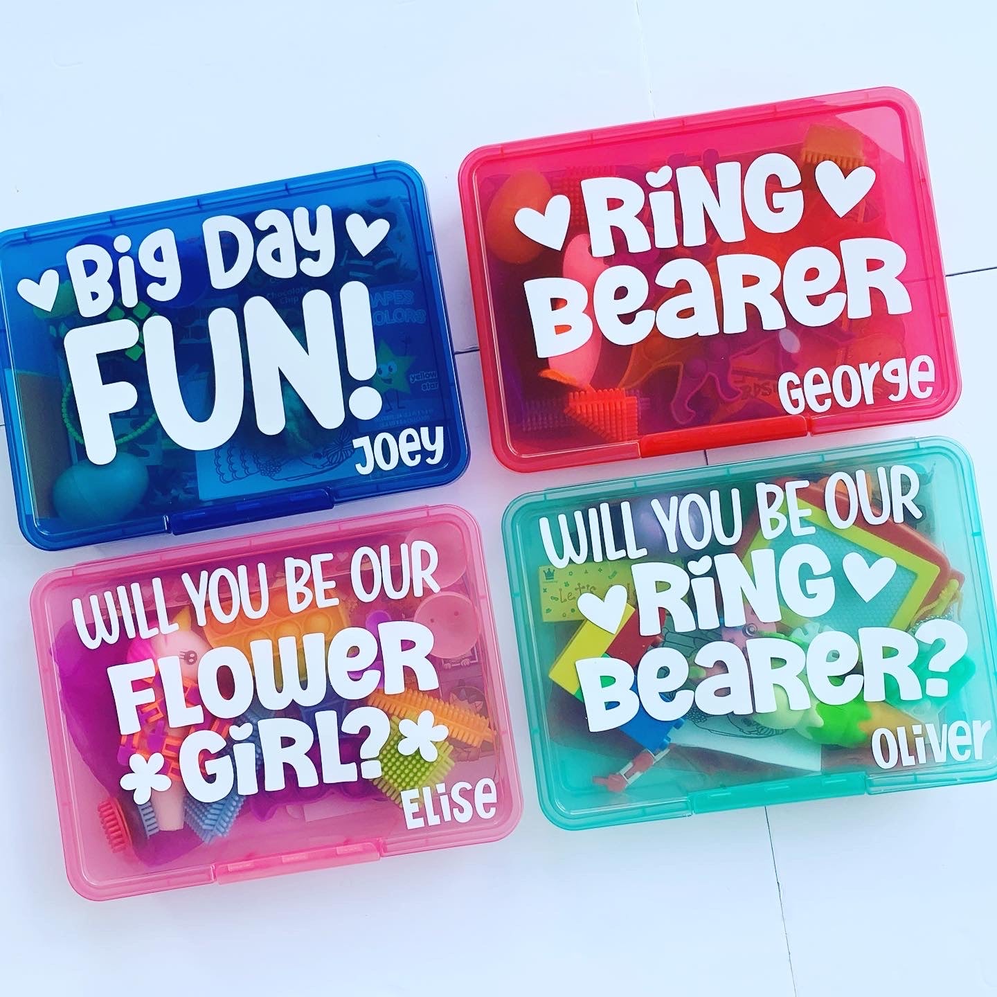 4 wedding themed busy boxes. Blue box with big day fun wording, 2 hearts and name Joey. Red box with ring bearer, 2 hearts and name George. Pink box with will you be our flower girl?, 2 flowers and name Elise. Turquoise ring bearer box name Oliver.