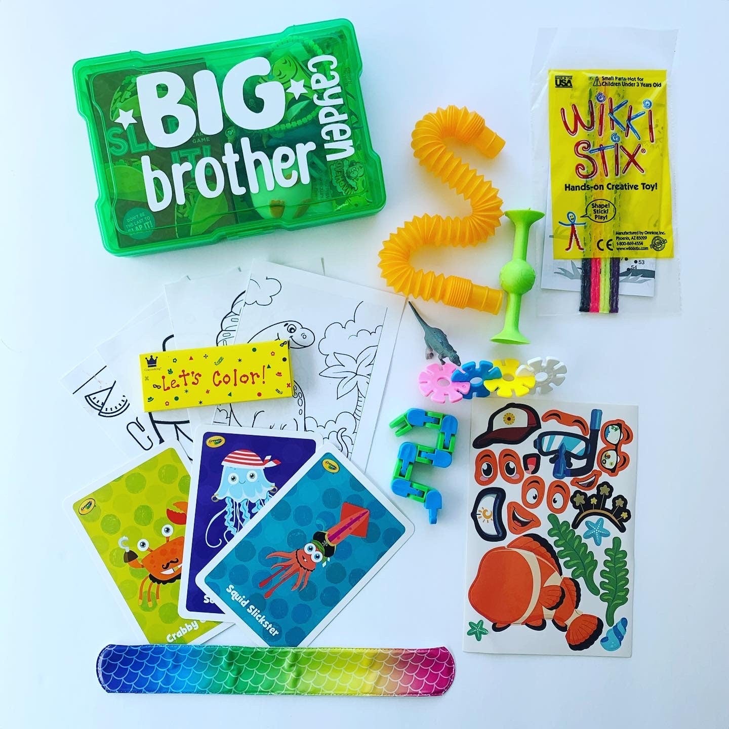 Green big brother busy box with the name Cayden personalized. Activities that come inside the box surround it. Pop tube fidget, slap bracelet, crayons, coloring pages, stickers, suction cup toy, Wikki Stix, animal matching cards.