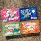 bright colored travel activity boxes - pink box with "up, up and away" with airplane, dark blue box with "road trip" and a sunshine, green box with "adventure awaits" and mountain, orange box with "are we there yet?" and arrow - all personalized with child's name