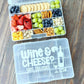 Snack container with fruits, crackers, and nuts on a wooden surface, featuring a clear lid with 'Wine & Cheese? Yes, please!' text.