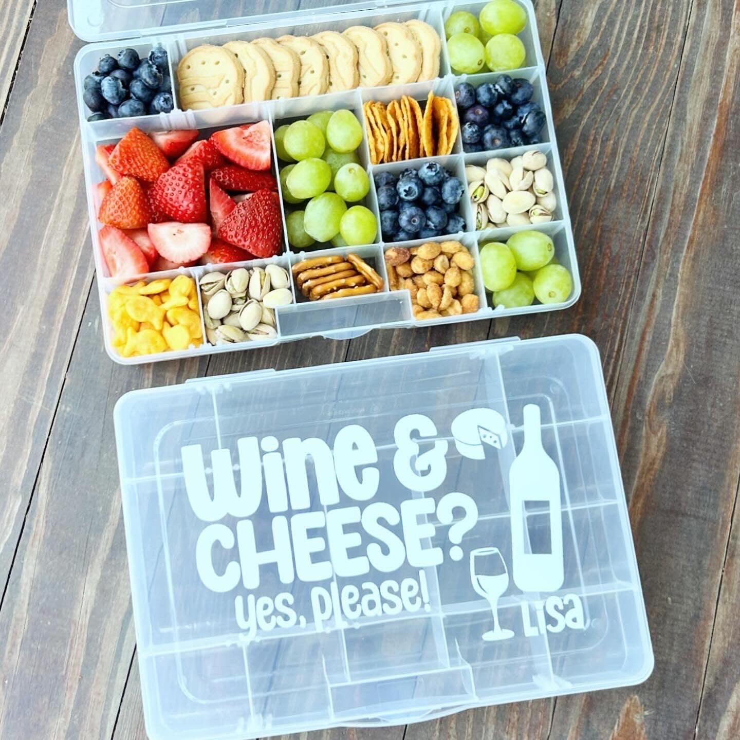 Snack container with fruits, crackers, and nuts on a wooden surface, featuring a clear lid with 'Wine & Cheese? Yes, please!' text.