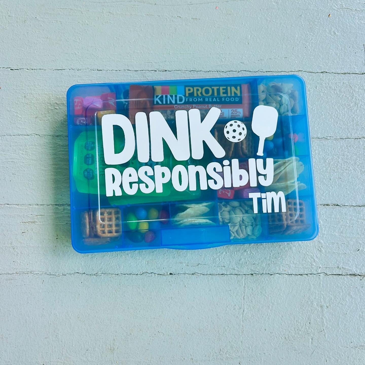 Blue snack container with 'Dink Responsibly Tim' text on a light blue wooden surface