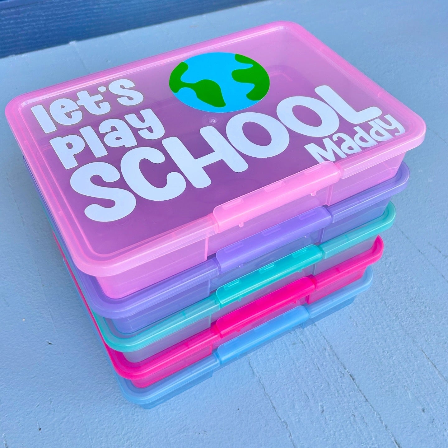 Stack of colorful school supplies containers with 'let's play school Maddy' text on a blue background