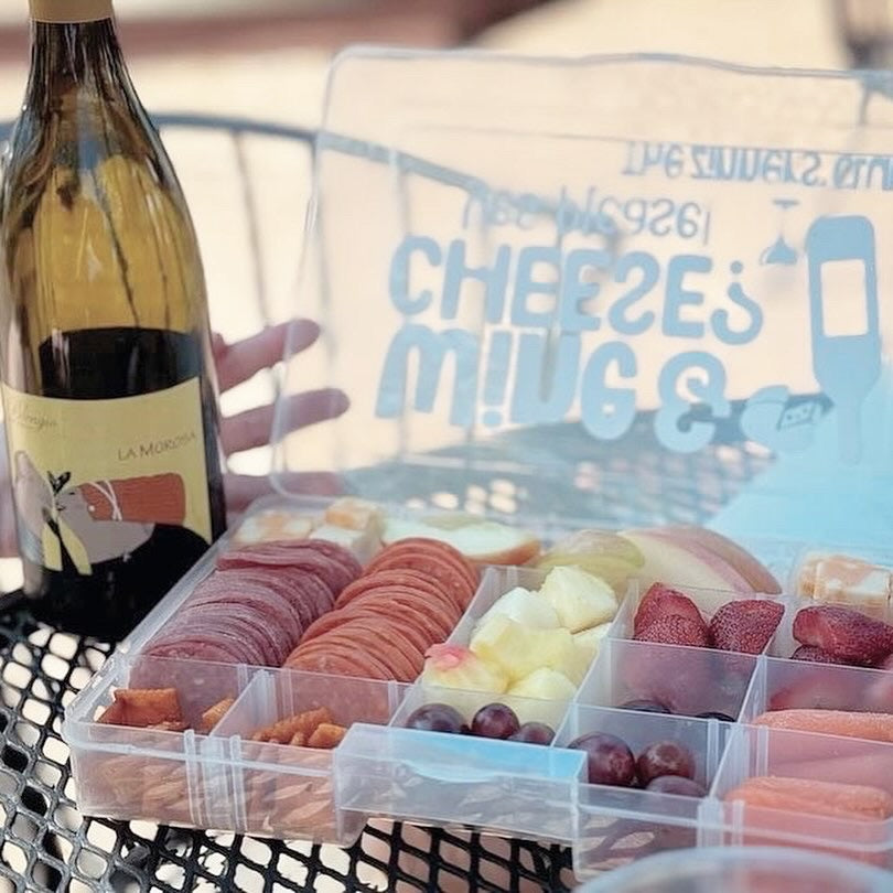 Transparent snack container with assorted foods next to a bottle of wine on a table.