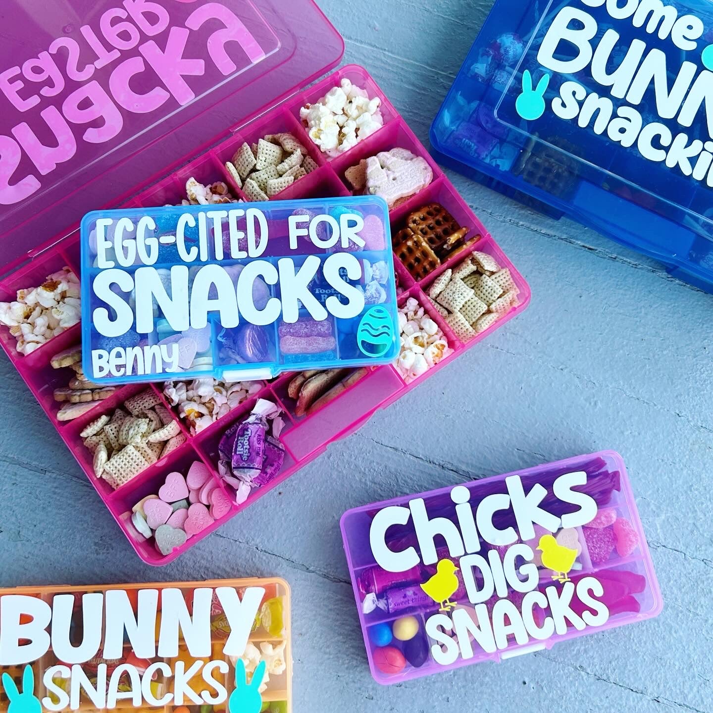 Pink snack box open showing compartments filled with various snacks. A smaller blue snack box on top of the pink one with egg cited for snacks, the name Benny and an easter egg on it. Also smaller boxes with bunny snacks, chicks dig snacks and some bunny snackin designs.