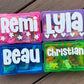 Four colorful name containers with names 'Remi', 'Lyla', 'Beau', and 'Christian' on a wooden surface.