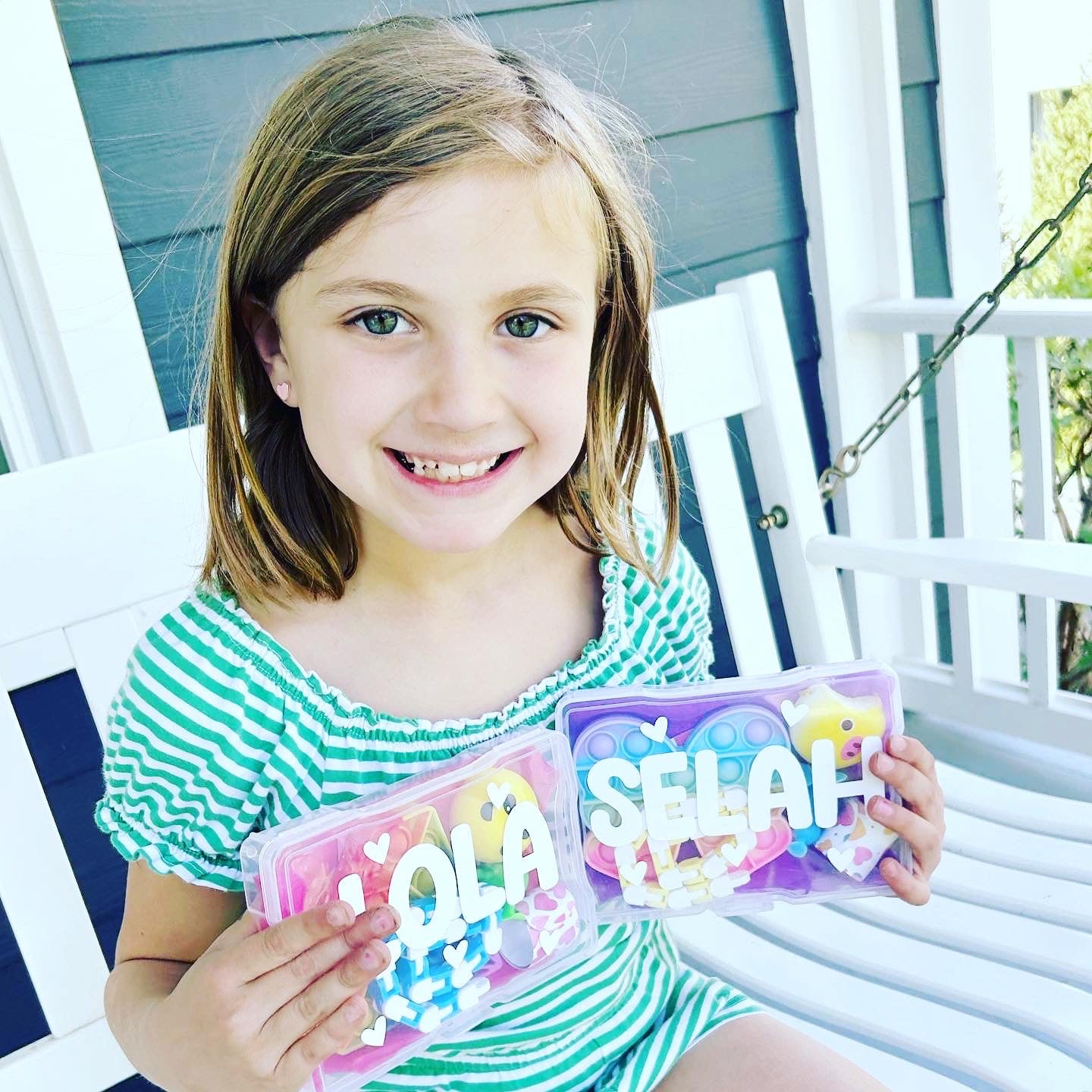 Young girl holding two clear plastic boxes with 'Lola and 'Selah' personalized on the front of the box. Each activity kit is filled with fun toys for children including a bendy snake fidget, slap bracelet, pop it fidgets and more.