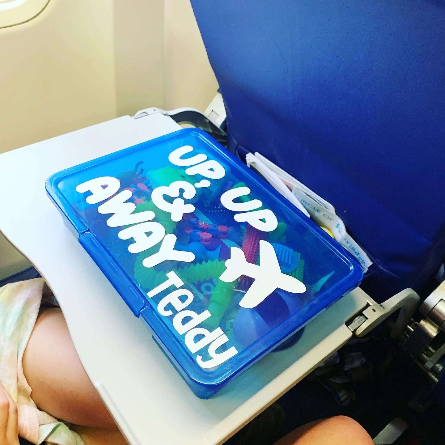 Dark blue travel activity box filled with kids toys with up, up and away, image of an airplane and personalized with the name Teddy on an airplane tray table