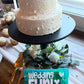 Two-tiered white wedding cake with decorative elements on a table, held by a person with a 'Wedding Fun! Teddy' sign.