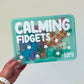 Teal box labeled 'Calming Fidgets' with various fidget toys inside, held by a hand.