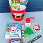 Christmas bucket with the name Benny on it which is filled with candy, pop it, coloring book, stickers and more. Contents are also laid out in front of the bucket.