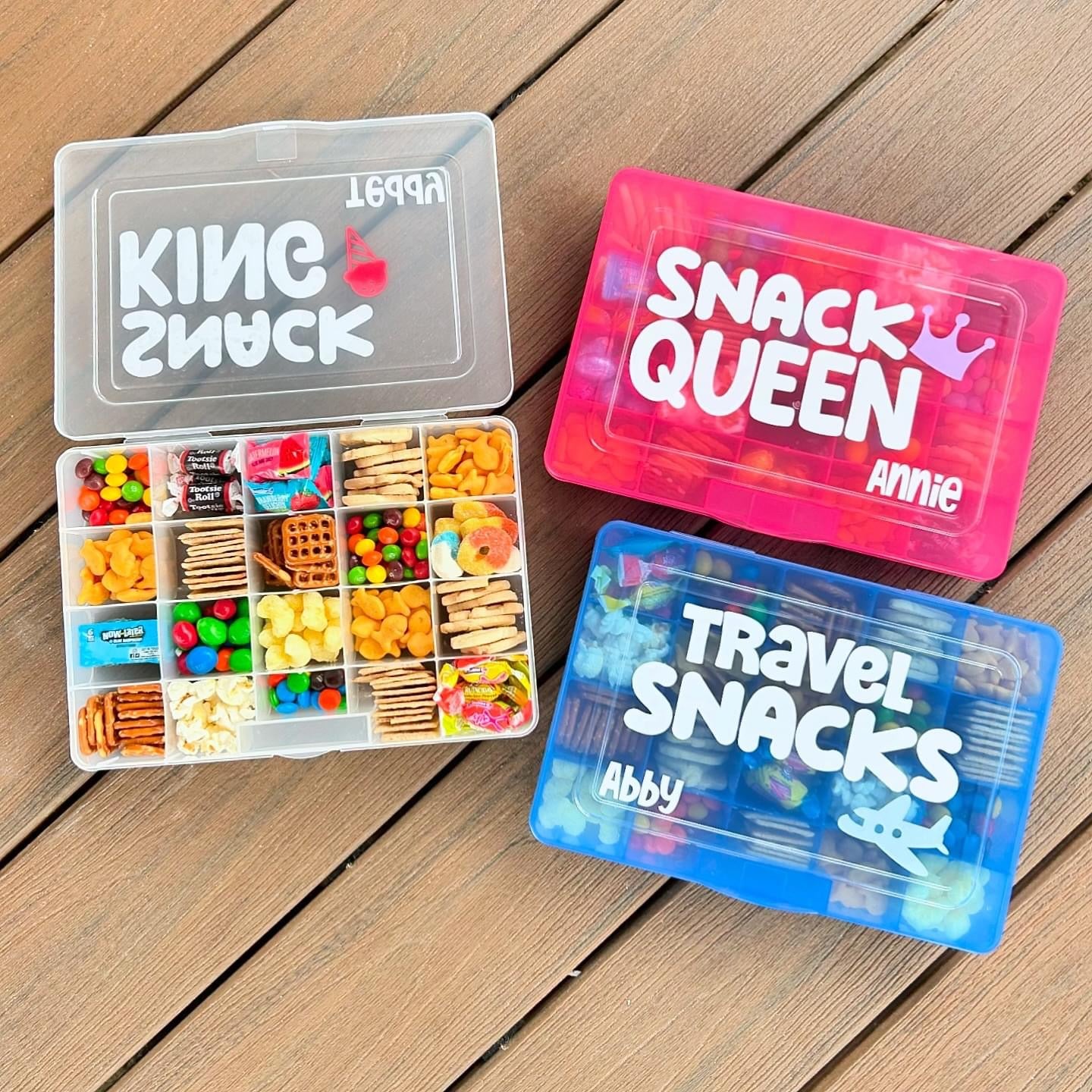 3 travel snack boxes. Clear box open with snacks inside each compartment. Goldfish crackers, skittles, tootsie rolls, pretzels, animal crackers, candy inside. Blue box with travel snack, name Abby and airplane. Pink Snack Queen box with crown.