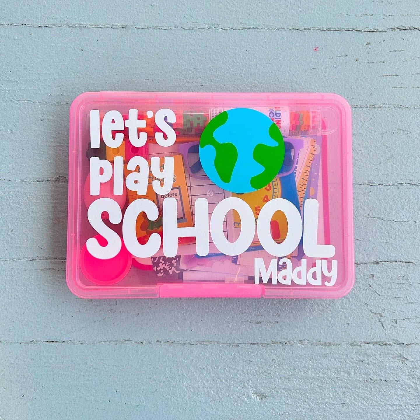 Pink toy storage box with 'Let's Play School Maddy' text on a light blue wooden surface