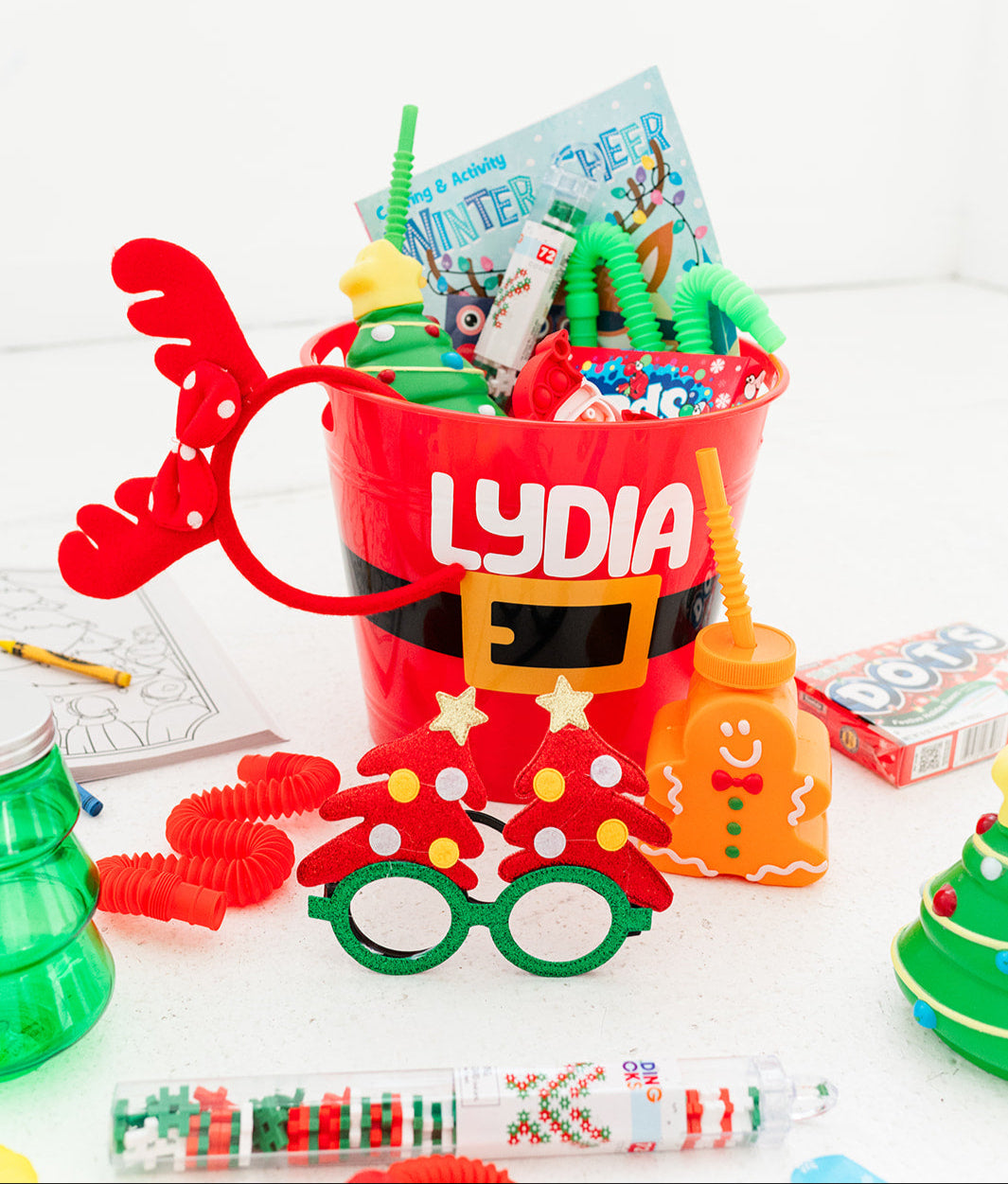 Red bucket with 'LYDIA' personalized on it, filled with children's toys and activities on a white background. Perfect for Christmas Eve or Christmas morning.