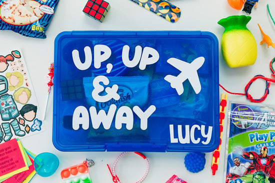 Blue toy box with 'Up, Up & Away Lucy' text surrounded by various toys and snacks on a white background