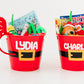 Two red Santa-themed buckets labeled 'Lydia' and 'Charlie' filled with toys on a white background.
