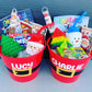 Two red Santa buckets serving as gift baskets, one labeled 'Lucy' and the other 'Charlie', filled with various Christmas-themed toys and items. Perfect for Christmas Eve or Christmas morning.