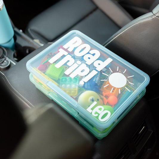 Two boxes are shown in the center console of a car. Top box has white lettering that says "Road Trip!" with a sunshine image and the name "Leo". The container is filled with various travel activities for kids such as fidgets, blocks, toys, snacks and more. Perfect for road trips or travel with toddlers and kids.