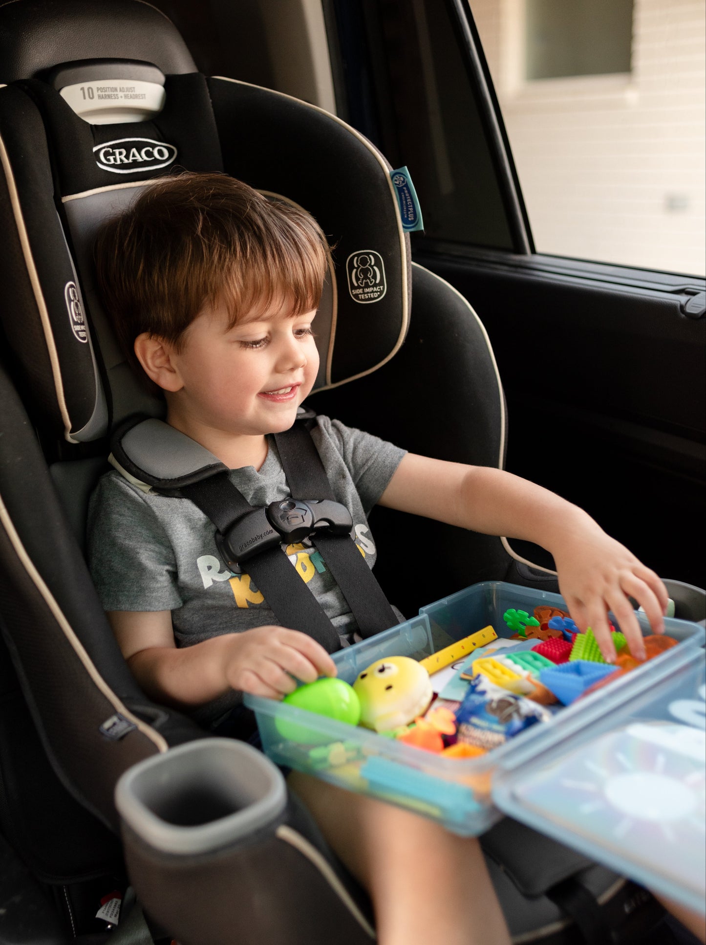 Child in a Graco car seat playing with a travel activity kit for kids. Blue plastic box is open in his lap with various toys and activities inside including fidgets, crayons, blocks, Oreo cookies, a green plastic egg, dinosaurs and more. 