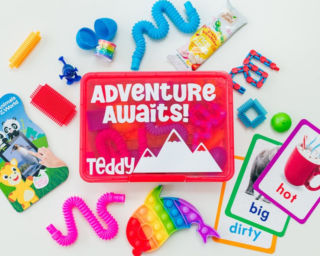 A red toddler travel busy box with the text 'ADVENTURE AWAITS' on the lid, surrounded by various colorful toys and activities including a peekaboo scarf, pop it fidget toy, building blocks, plastic eggs, a slap bracelet, pop tubes, a slow-rise squishy, and learning flashcards.
